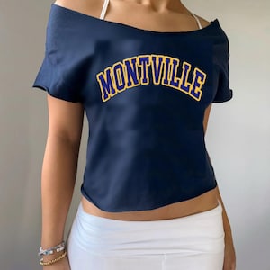 May include: A navy blue off-the-shoulder crop top with the word "MONTIVILLE" printed in yellow and blue letters.