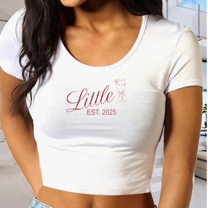 Puede incluir: Top corto blanco con cuello redondo. La camiseta presenta la palabra "Little" en cursiva, junto con "EST. 2025" y un gráfico de copa de vino en rojo. La camiseta está hecha de un material suave y cómodo.
