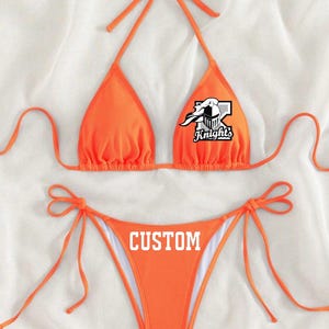 May include: Orange bikini top and bottom with a white graphic of a knight's helmet and the word "Knights". The bottom has the word "CUSTOM" printed on it.