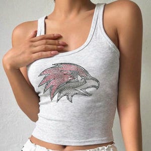 May include: A light gray ribbed tank top featuring a rhinestone eagle design in black and red. The sleeveless top has a scoop neck and a cropped fit, perfect for casual wear.
