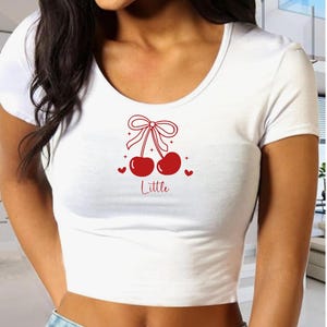 Puede incluir: Top corto blanco con cuello redondo y mangas cortas. La camiseta presenta un diseño de cerezas rojas con un lazo, corazones y la palabra "Little" impresa debajo.