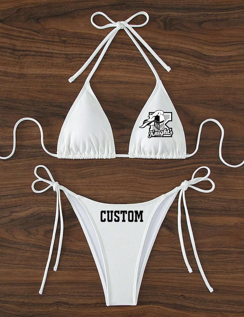 May include: White bikini with a custom design. The top has a black and white graphic of a knight's helmet with the word "Knights" below it. The bottom has the word "CUSTOM" printed in black.