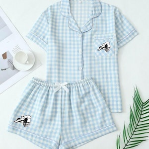 May include: Light blue and white checkered pajama set. The set includes a short-sleeved button-up shirt with a chest pocket and matching shorts. Both pieces feature a black and white graphic. The shorts have a white bow at the waist.