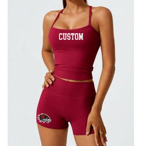 May include: A maroon athletic set featuring a sports bra and shorts. The sports bra has thin straps and the word "CUSTOM" in white block letters. The shorts have a logo on the left leg. This activewear set is designed for workouts.