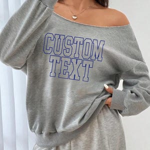 May include: Gray off-the-shoulder sweatshirt with blue outlined text that says "CUSTOM TEXT".