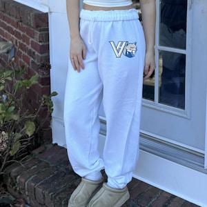 May include: White sweatpants with a drawstring waist and elastic cuffs. The pants feature a blue and white logo on the left leg. The person is wearing light brown slippers.