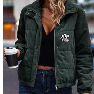 May include: Dark green corduroy puffer jacket with a stand-up collar, zip closure, and a small logo that says "Knight's" on the chest. The jacket has a quilted design and is worn with a black top and blue jeans.
