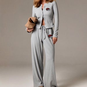 May include: Light gray loungewear set featuring a button-down cardigan and wide-leg pants. The set includes a drawstring waist and a small embroidered emblem on the cardigan and pants. The model is wearing the set with brown shoes.