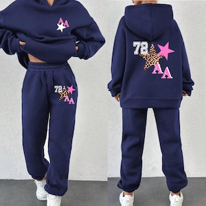 May include: A navy blue two-piece tracksuit with a hoodie and sweatpants. The hoodie has a large pink star with a leopard print and the letters "AA" and the number "72" on the back. The sweatpants have a smaller pink star with a leopard print and the letters "AA" and the number "78" on the left leg.