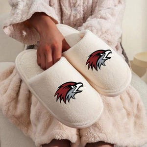 May include: A pair of cream-colored, plush slippers featuring a maroon and gray eagle logo on the top. The slippers are soft and cozy, perfect for indoor wear. The eagle logo adds a touch of collegiate or team spirit.