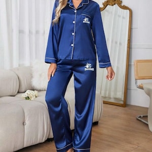 May include: Navy blue satin pajama set with white piping. The long-sleeved top has a button-down front and a Sharks logo on the chest pocket. The matching pants are wide-legged. The set is designed for comfort and relaxation.