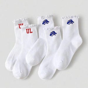 May include: White crew socks with ruffled tops. Two pairs feature the letters "UL" in red, while the other two pairs display a blue lion logo. The socks are made of a ribbed knit material.