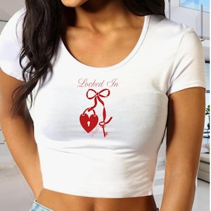 Puede incluir: Top corto blanco con cuello redondo y diseño de cerradura y llave en forma de corazón rojo. Las palabras "Locked In" están encima de la cerradura y la palabra "Little" debajo. La parte superior es de material ligero.