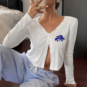 May include: White long-sleeve cardigan with a V-neck and lace trim. The cardigan has a blue emblem on the chest and is buttoned down the front. The model is wearing blue and white striped pants.
