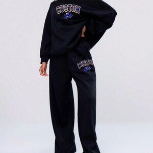 May include: Black off-the-shoulder sweatshirt and matching wide-leg sweatpants. The word "CUSTOM" is printed in gold with a blue and white graphic. The same design is repeated on the sweatpants. A casual, comfortable outfit.