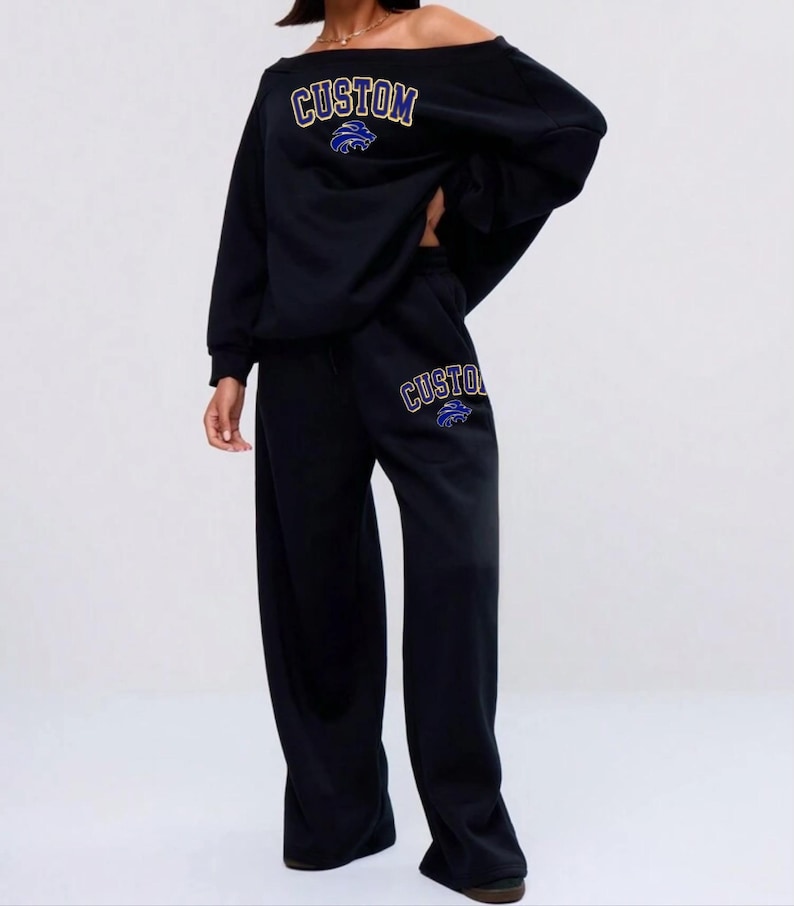 May include: Black off-the-shoulder sweatshirt and matching wide-leg sweatpants. The word "CUSTOM" is printed in gold with a blue and white graphic. The same design is repeated on the sweatpants. A casual, comfortable outfit.
