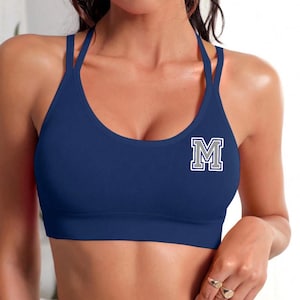 Puede incluir: Un sujetador deportivo azul marino con un diseño de doble tirante. El sujetador presenta la letra "M" blanca y gris en la parte delantera. Diseñado para actividades deportivas.