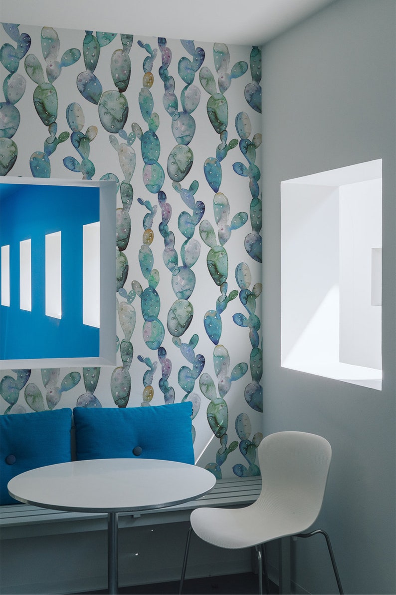 May include: A white room with a cactus patterned wallpaper, a blue bench with two blue cushions, a white round table, and a white chair. The room has a window with a white frame.