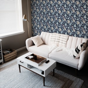 May include: A white couch with a white throw blanket and a black and white patterned pillow. The couch is in a living room with a white coffee table, a white end table, and a record player on a white shelf. The walls are covered in a blue and white floral wallpaper.