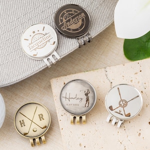 Könnte beinhalten: Eine Sammlung personalisierter Golfballmarker in verschiedenen metallischen Ausführungen. Designs umfassen gekreuzte Golfschläger, Initialen, Namen und Daten. Ein Marker ist an einer grauen Golfmütze befestigt, ein weißer Golfball ist ebenfalls sichtbar.