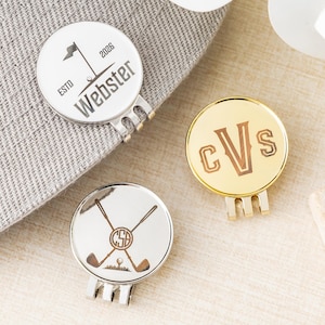 Könnte beinhalten: Drei Golfballmarker mit verschiedenen Designs. Einer ist silberfarben mit einer Flagge und dem Namen "Webster". Ein anderer ist goldfarben mit den Initialen "CVS". Der dritte ist silberfarben mit gekreuzten Golfschlägern und einem Golfball.