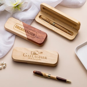 Op de afbeelding: Houten pennenkokers en pennen met gepersonaliseerde gravures. Eén koker is open en onthult een pen. De kokers zijn gegraveerd met de namen "ENCARNACION" en "Gallagher". De pennen zijn bruin en goud.