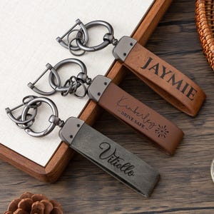 May include: Three personalized leather keychains with metal hardware. The keychains are brown, gray, and tan, each with a name or message engraved. One says "JAYMIE", another "Kimberley - DRIVE SAFE", and the third "Vitiello".