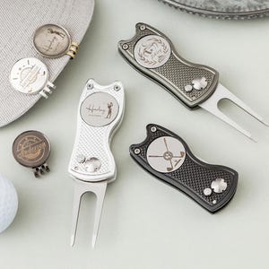 May include: A collection of golf accessories, including a white golf ball, personalised ball markers, and divot tools. The ball markers are silver and bronze with engraved names. The divot tools are silver and black with a textured grip.