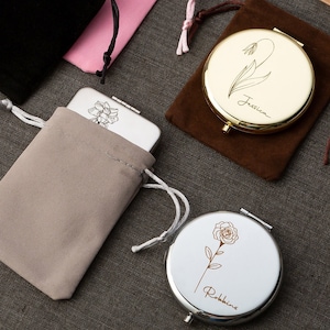 Puede incluir: Dos espejos compactos con diseños florales y nombres personalizados. El espejo de la izquierda tiene un acabado plateado con un diseño de narciso y el nombre "Jessica". El espejo de la derecha tiene un acabado dorado con un diseño de rosa y el nombre "Robbins".