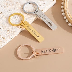 Puede incluir: Tres llaveros dorados con etiquetas grabadas personalizadas. El primer llavero tiene el nombre "Sehan" grabado con una corona. El segundo llavero tiene el nombre "Manuel Alahimi" grabado con un corazón. El tercer llavero tiene el nombre "ALEX" grabado con una huella de pata.