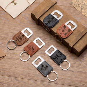 May include: A collection of personalized bottle opener keychains in various colors, including brown, black, and gray. Each keychain features a metal bottle opener and a leatherette tag with custom engravings, such as names and initials. The keychains are arranged on a wooden surface.