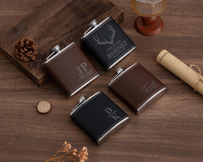 May include: Four personalized flasks with silver accents and leather-like covers are arranged on a wooden surface. The flasks are brown and black, with custom text and designs, including initials, names, and a deer antler graphic. A pine cone and scroll are also visible.