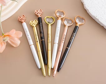 May include: Seven gold and rose gold heart-shaped pens with personalized names. The pens have clear crystal accents and are arranged on a light brown surface.