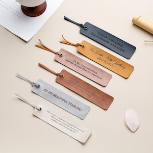 Può includere: Cinque segnalibri in pelle di diversi colori con testo inciso. Il testo include citazioni, un messaggio di compleanno e un messaggio personale. I segnalibri sono su una superficie beige chiaro.