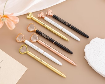 May include: An assortment of elegant pens with heart-shaped toppers. The pens feature gold, rose gold, and black barrels, with white accents. Some pens have names engraved on them. The pens are arranged on a neutral surface.