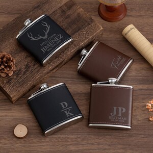 May include: Four personalized flasks in black and brown leather with silver accents. The flasks have custom engravings, including names, initials, and the text "Best Man." The flasks are displayed on a wooden surface.