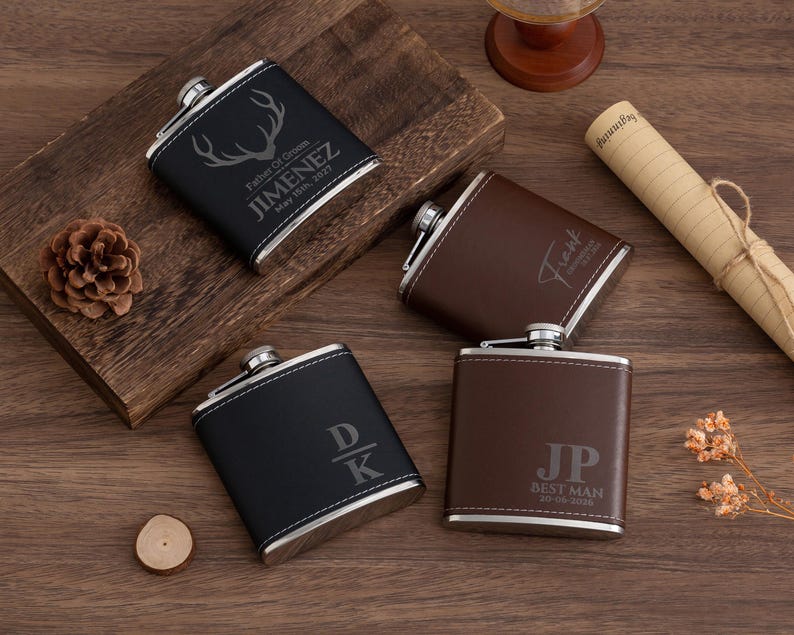 May include: Four personalized flasks in black and brown leather with silver accents. The flasks have custom engravings, including names, initials, and the text "Best Man." The flasks are displayed on a wooden surface.