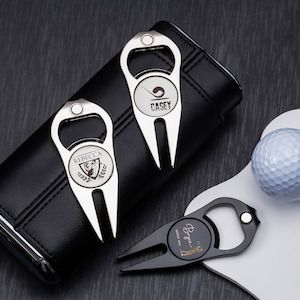 Op de afbeelding: Een verzameling golfaccessoires, waaronder twee zilveren flesopener-divotgereedschappen met gepersonaliseerde gravures, een zwart divotgereedschap, een golfbal en een zwart leren etui. De gereedschappen hebben een flesopenerfunctie.