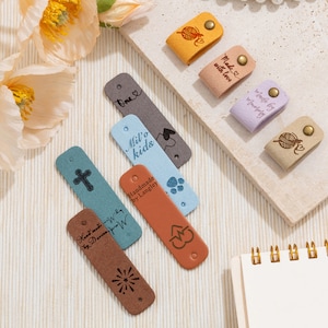 May include: A selection of leather tags in shades of brown, beige, blue, and lilac. The tags display various designs and text, including a cross, paw prints, and a heart. Some tags have the text "Handmade by Langley" and "Made with love."