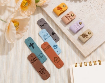 May include: A selection of leather tags in shades of brown, beige, blue, and lilac. The tags display various designs and text, including a cross, paw prints, and a heart. Some tags have the text "Handmade by Langley" and "Made with love."