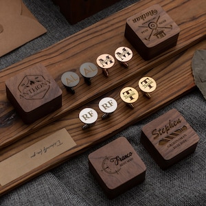 May include: A set of personalized cufflinks in various colors and metals, including gold, silver, and rose gold. Each cufflink features a different initial and is displayed in a wooden box with a personalized inscription. The boxes are made of dark wood and have a compass design.