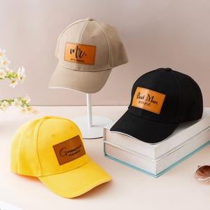 Op de afbeelding: Drie baseballpetten in beige, zwart en geel. Elke pet heeft een leren patch met de tekst "Mr.", "Best Man" en "Groomsmann". De petten worden gepresenteerd op een tafel met een vaas bloemen en een zonnebril.
