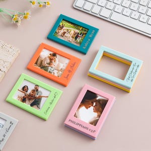 Pode incluir: Uma coleção de molduras magnéticas retangulares e coloridas em azul-petróleo, laranja, verde-claro, rosa e cinza. Cada moldura apresenta texto gravado personalizado, incluindo nomes e datas, e contém uma fotografia ou está vazia.