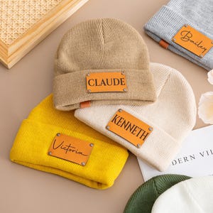 Puede incluir: Varios gorros personalizados en colores como beige, amarillo, gris y crema. Cada gorro tiene un parche rectangular de cuero con un nombre. Los nombres son "Claude", "Victoria", "Kenneth" y "Bailey".