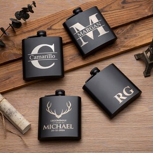 May include: Four black metal hip flasks with personalised engravings. The flasks feature different designs, including initials, names, and a stag silhouette. The flasks are arranged on a wooden surface.