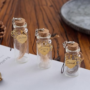 May include: Three small glass bottles with cork stoppers, each containing a lock of hair. Gold-colored heart-shaped charms with names hang from the bottles. The bottles are on a white surface with a wooden background.