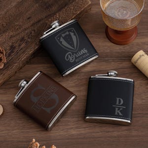 May include: Three personalized flasks in black and brown leather with silver accents. One flask has a tuxedo graphic and the words "Bruns Best Man". Another has the initial "S" and the last name "Hopkins". The third flask has the initials "DK".