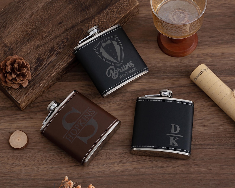 May include: Three personalized flasks in black and brown leather with silver accents. One flask has a tuxedo graphic and the words "Bruns Best Man". Another has the initial "S" and the last name "Hopkins". The third flask has the initials "DK".