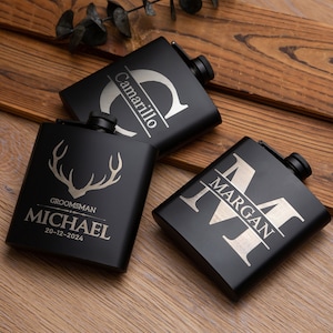 Könnte beinhalten: Drei schwarze Metall-Schnapsflaschen mit gravierten Designs. Die erste Flasche trägt den Namen "Camarillo" und einen gravierten Buchstaben "C". Die zweite Flasche hat ein Hirschgeweih und den Text "Groomsman Michael 20-12-2024". Die dritte Flasche trägt den Namen "Margan" und den gravierten Buchstaben "M".