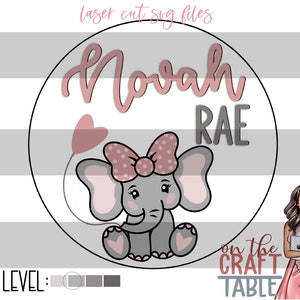 Puede incluir: Un círculo rosa y gris con el nombre "Nova Rae" en escritura rosa y un elefante de dibujos animados sosteniendo un globo en forma de corazón rosa. El elefante tiene un lazo rosa con lunares blancos. El texto "on the craft table" está debajo del círculo.